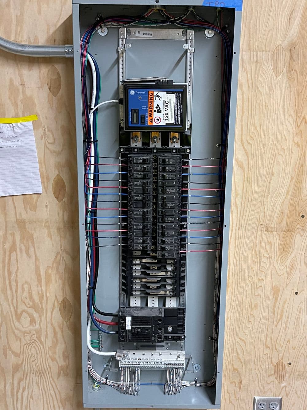 Electrical panel with circuit breakers, wires, and safety labels mounted on a wooden wall.