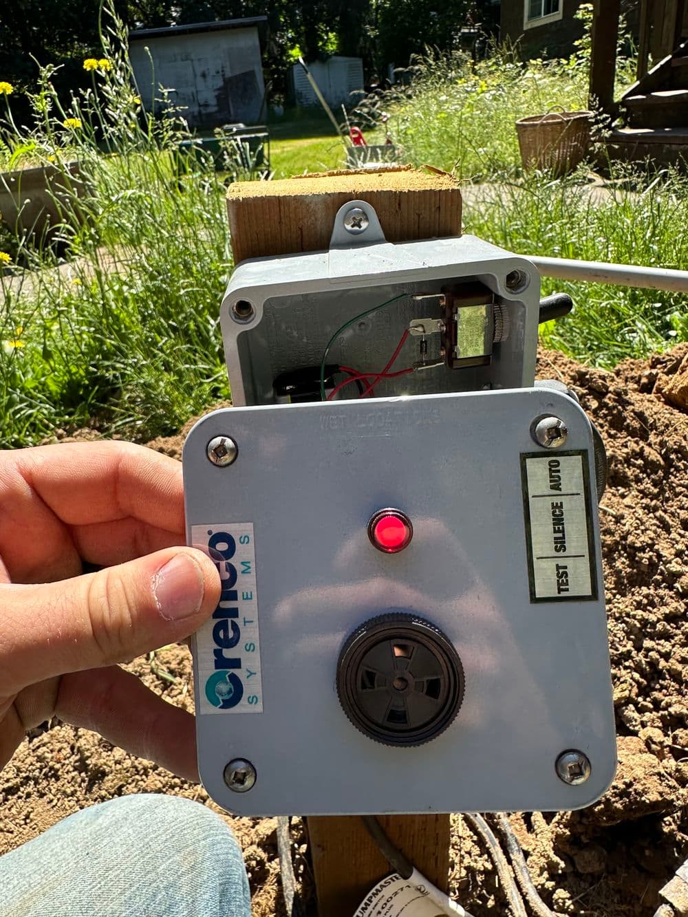 Hand holding a gray control box with a red light, part of a irrigation system installation.